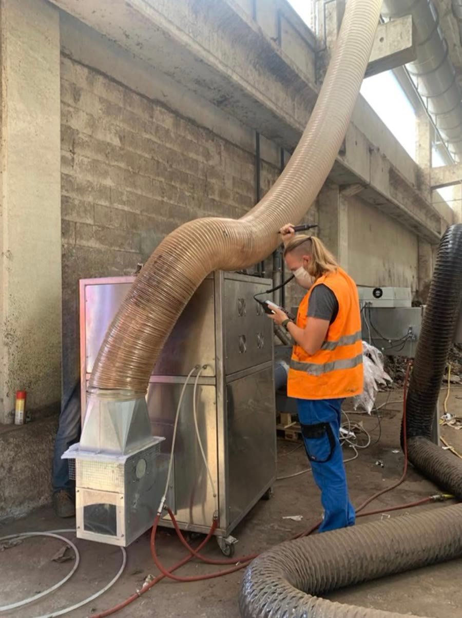 A worker in high visibility clothing taking a reading with a handheld instrument beside ducting on an industrial site